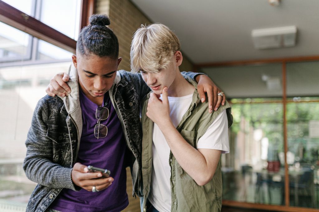 Two high school friends looking at pictures on a smartphone together with theirs arms around each others shoulders.