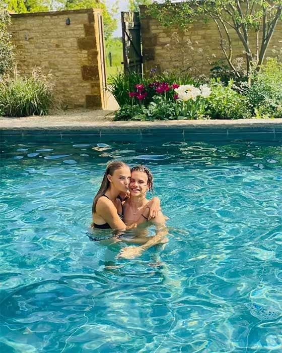a teenage couple embrace in an outdoor pool on a sunny day with a walled garden in the background