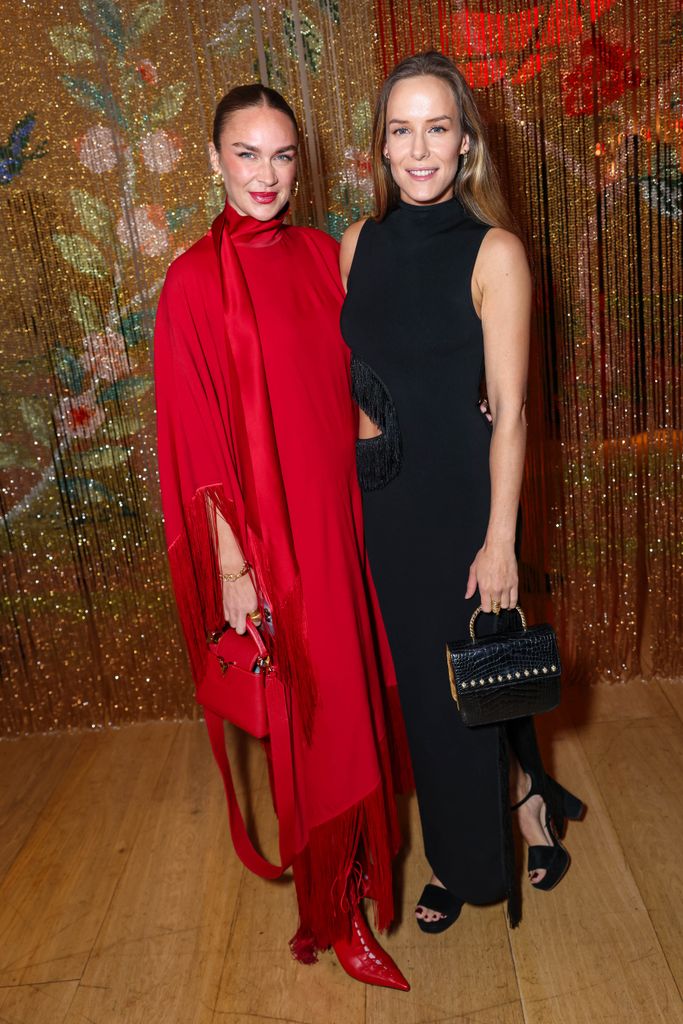 Nina Suess and Hum Fleming stand against a shimmering gold backdrop. One wears a bold red fringe cape dress with matching heels, while the other stuns in a sleek black dress with a textured finish. The scene exudes pure festive glamour.