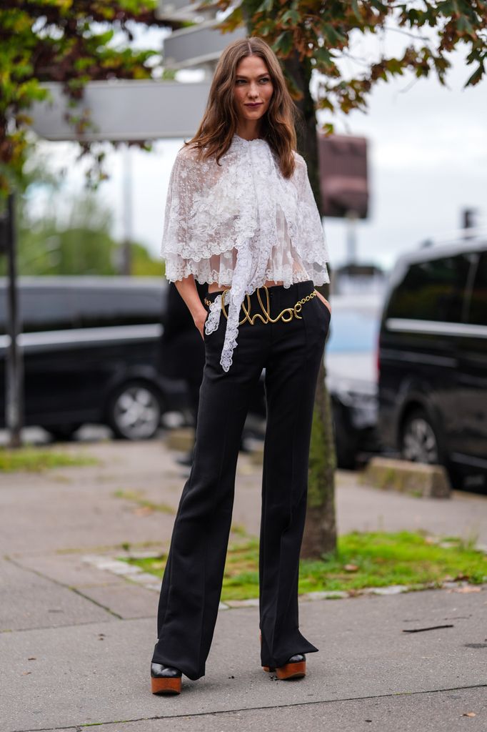 Karlie Kloss wears white sheer mesh lace flowy layered shirt, black high low-rise pants, gold Chloe belt, shiny black platform brown Chloe heels, outside Chloe, during the Paris Fashion Week Spring/Summer 2025
