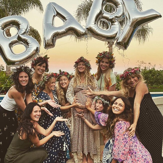 a group of women wearing flower garlands on their heads gather around a heavy pregnant meredith and all of them look at the camera while each extending a hand out to touch her baby bump and goldie is the furthest away but stretches to reach as the sun sets in the background and a helium balloon reading baby is visible over their heads