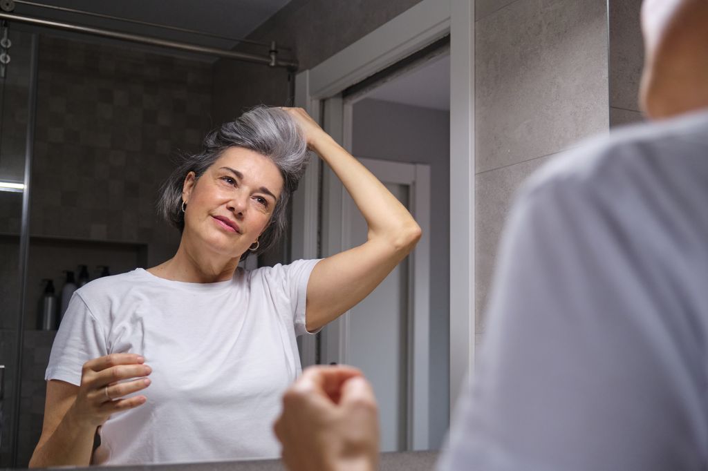 Mature woman taking care of her gray hair, styling it in front of the bathroom mirror