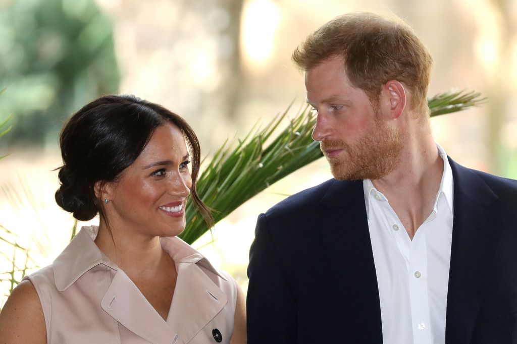 Prince Harry in a suit looking at Meghan Markle in front of a palm tree