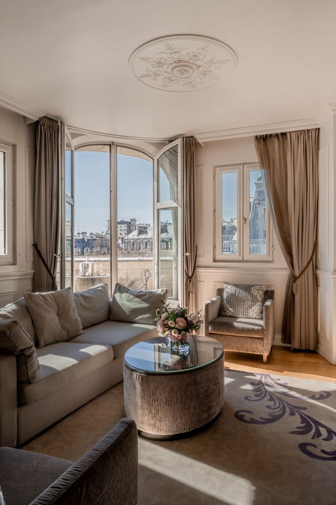 Interior of a luxury suite at La Clef Louvre Paris, featuring sunlit French windows overlooking Paris rooftops, a stylish grey sofa, and elegant classical wall moldings.