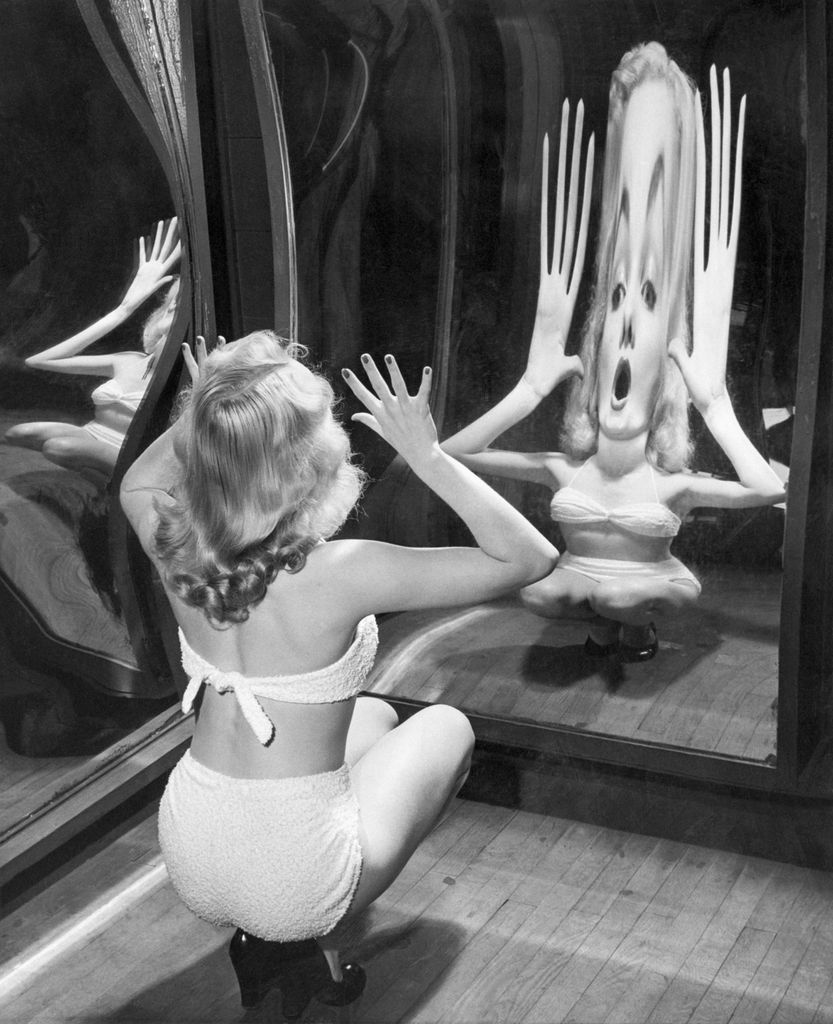Nothing that Alice ever found behind the looking-glass could be half as weird as the reflection produced by blonde Canda Loden as she tests one of the new trick mirrors at the Rockaways Playland. | Location: Queens Amusement Park, Borough of Queens, New York, USA.