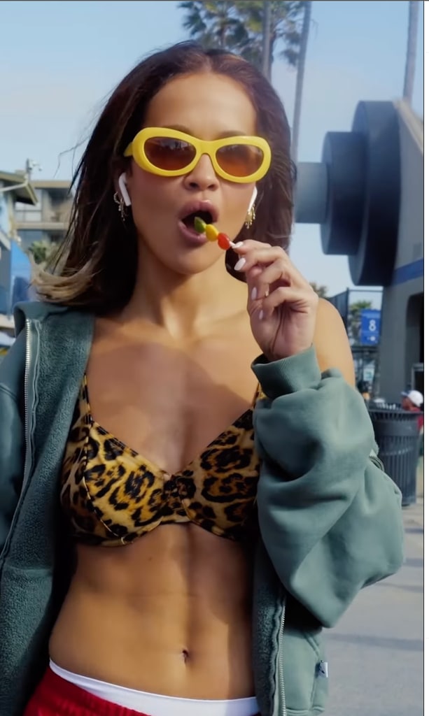 Rita Ora wears a leopard print bikini top, grey/green hoodie and bright yellow sunglasses. She sucks on a lollipop and showcases her six pack. 