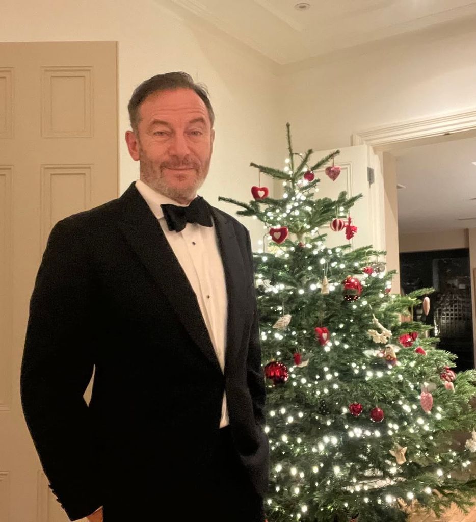 Jason Isaacs wearing a tuxedo standing by a Christmas tree in living room at home
