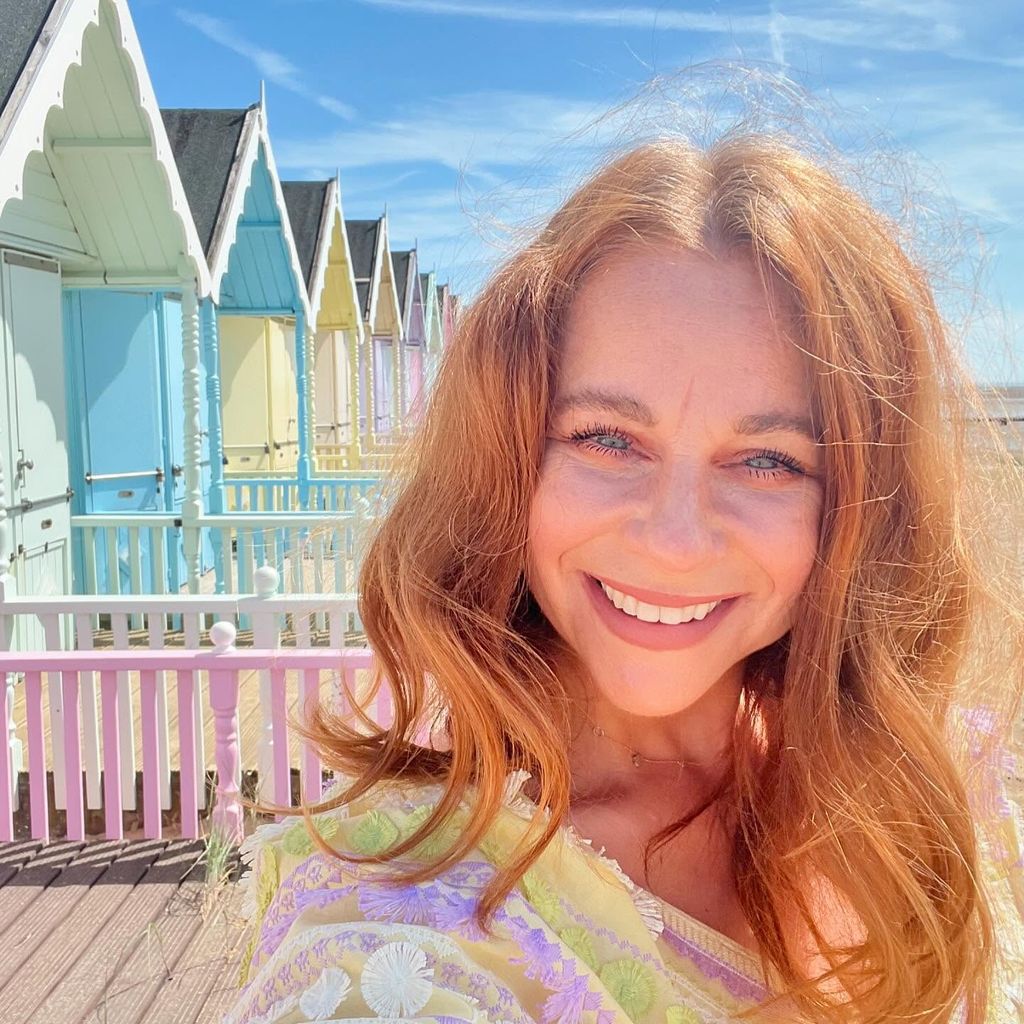 Headshot of a redheaded woman smiling in the sun in front of pastel beach huts