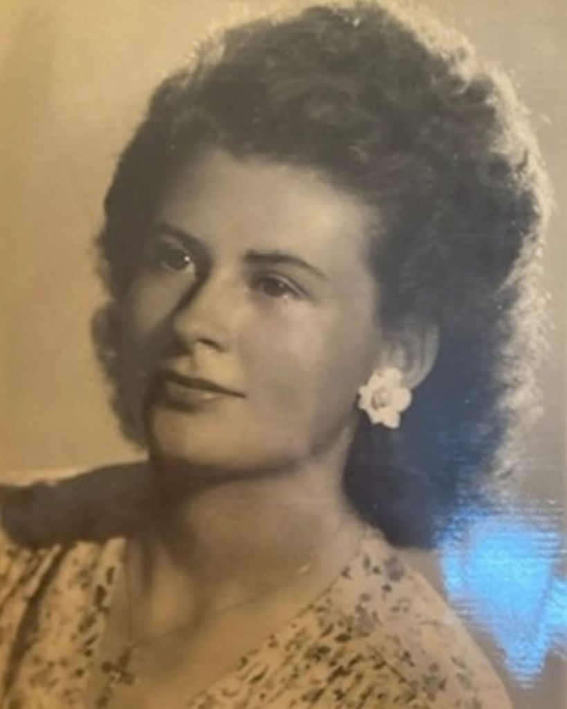 A vintage studio portrait of Lily James' French grandmother wearing a delicate floral dress and white flower earrings. Her soft expression and voluminous curls reflect mid-century femininity and the timeless beauty that clearly runs in the family.