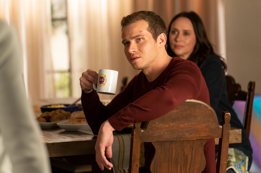 Oliver Stark holds a mug as he sits at a dining table in a still from 911
