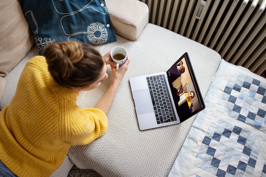 Directly above shot of a woman lying on couch with coffee cup and video calling her friends on laptop. Young female video chatting her friends while lying on sofa at home.
