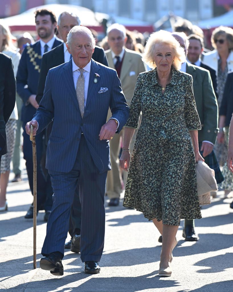 King Charles III walking at Chelsea Flower Show 2025 wearing a navy pinstripe suit, lilac tie, and carrying a wooden cane, next to Queen Consort Camilla in green floral dress.