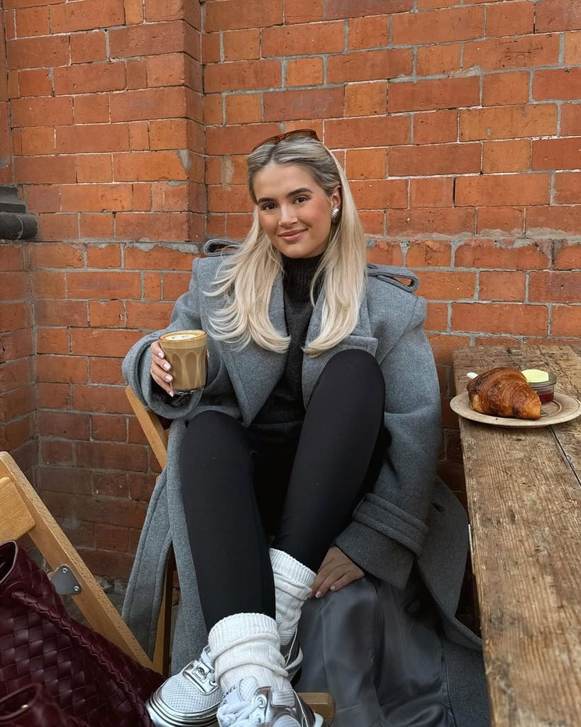 Molly-Mae sits at a cozy outdoor café table, wearing a grey belted wool coat, black leggings, and sneakers. She smiles while enjoying coffee and a croissant, embodying relaxed elegance.