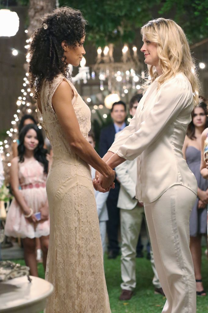 Stef et Lena de The Fosters se font face, se tenant la main. Lena porte une robe longue champagne et Stef porte un costume blanc.