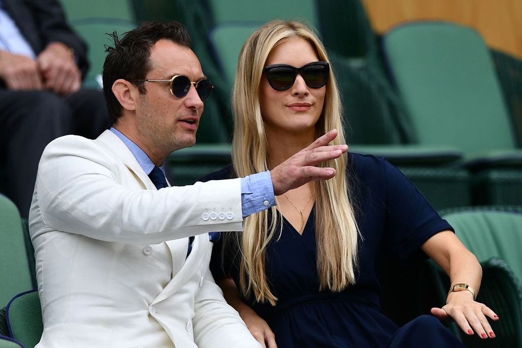 Jude Law and his wife Phillipa Coan sat at Wimbledon, both wearing sunglasses and looking to the left, Jude is pointing out to the left