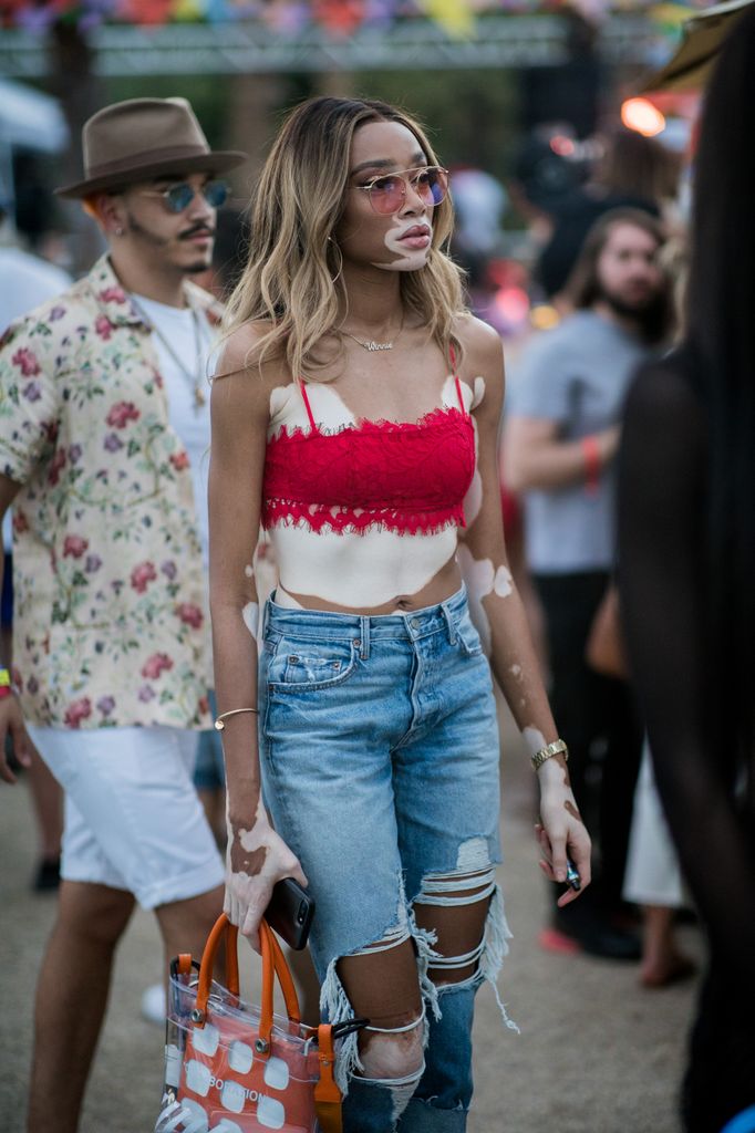 Winnie Harlow wears a red lace crop top and ripped jeans at Coachella, accessorizing with pink sunglasses and a clear polka-dot tote.