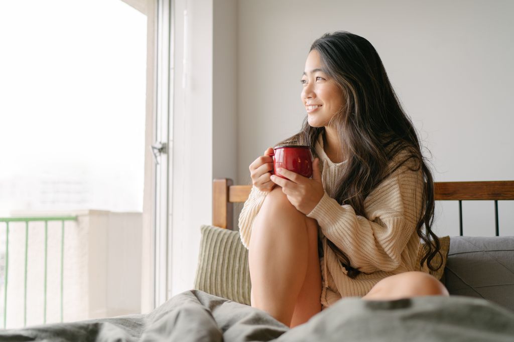 A beautiful young woman is drinking a cup of hot drink in her bed and looking out of the widow in the morning.