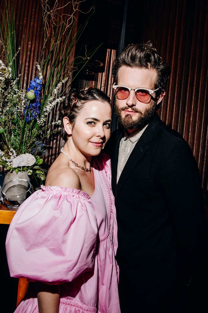 Bethany C. Meyers wearing a pink off-shoulder dress and Nico Tortorella in a black suit wearing sunglasses
