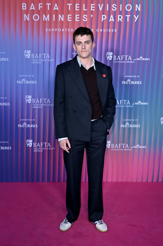 Jake Dunn wears a black blazer and trousers, white trainers, jumper and shirt on the pink carpet