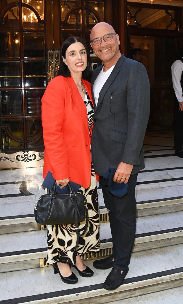 Gregg Wallace in a grey suit smiling with his wife in a black and white jumpsuit and red blazer