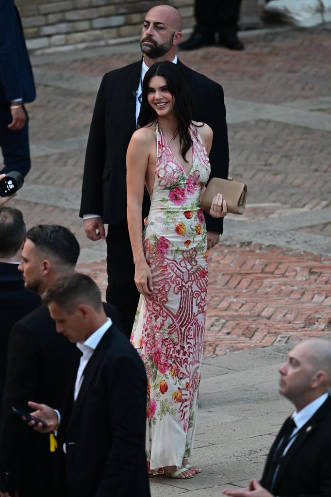 Kendall Jenner walks through Venice in a floral halterneck gown with pink embroidery and lace detail, styled with a nude clutch and beachy waves.