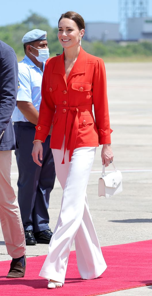 Kate Middleton in red jacket and white trousers carrying a white handbag