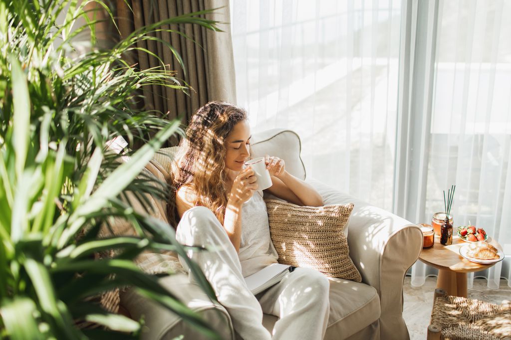 Beautiful woman have relax at home with cup of tea and book. Young woman at home sitting on modern chair and relaxing in her living room reading book and drinking coffee or tea. Reading activity for adult.