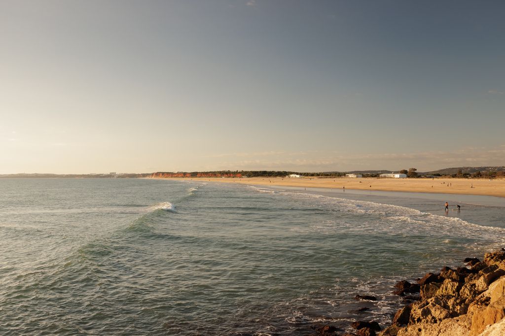 Golden hour view of Rocha Baixinha Beach in Vilamoura, Portugal, with gentle waves rolling onto the sandy shore and cliffs in the distance.