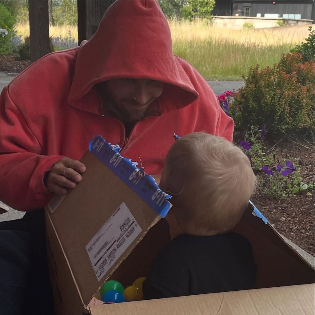photo of justin bieber wearing red hoodie smiling at son jack who is sitting in a cardboard box with his back to the camera