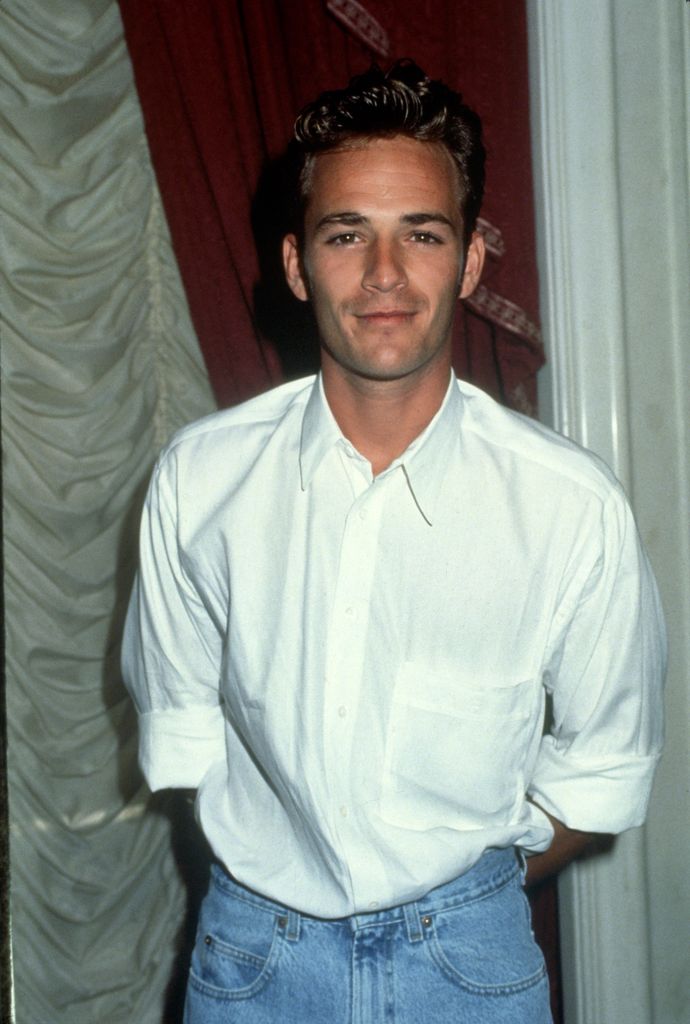 Luke Perry porte une chemise blanche et un jean, les mains dans les poches arrière.
