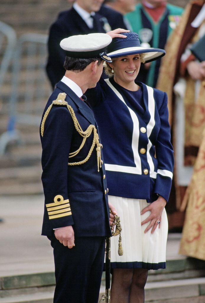 The late Princess Of Wales in navy and white dress holding onto hat beside prince charles in navy uniform