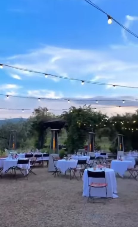 Outdoor event setup at dusk with round tables covered in white cloths, decorated with centerpieces, surrounded by chairs. String lights hang overhead, and there are patio heaters among the tables. Trees and hills are visible in the background.