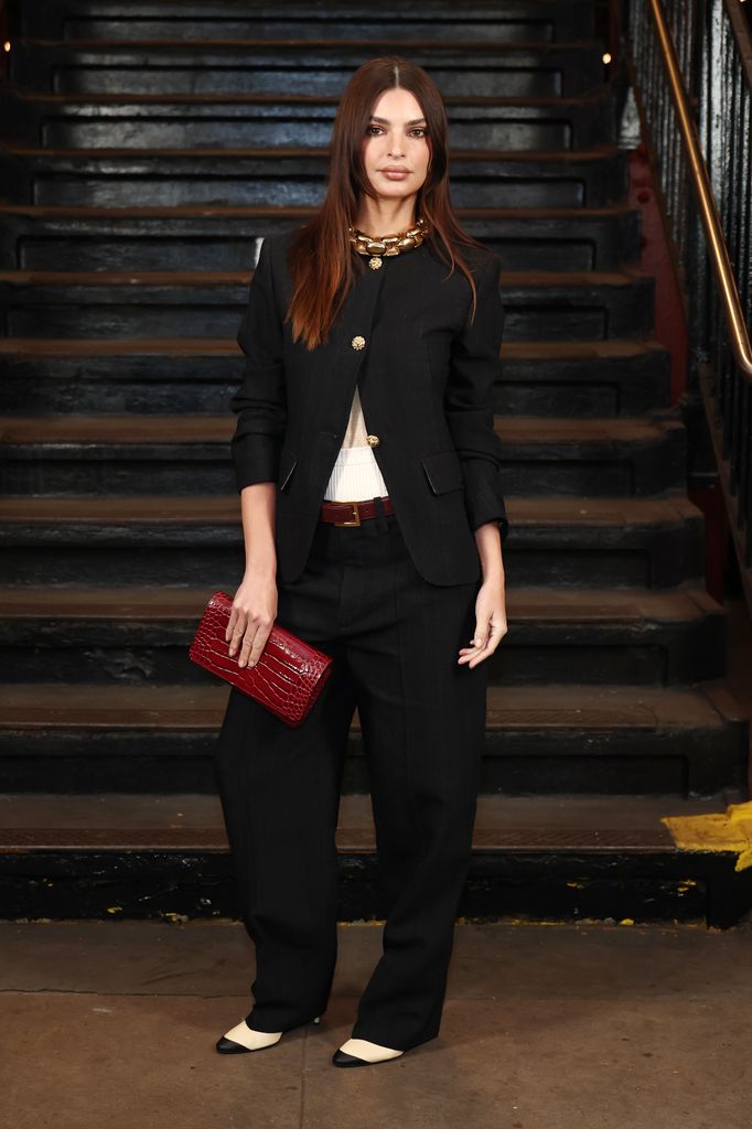 Emily Ratajkowski stands at the foot of the subway stairs, wearing a black blazer with gold buttons, matching trousers, monochrome heels and a red clutch bag