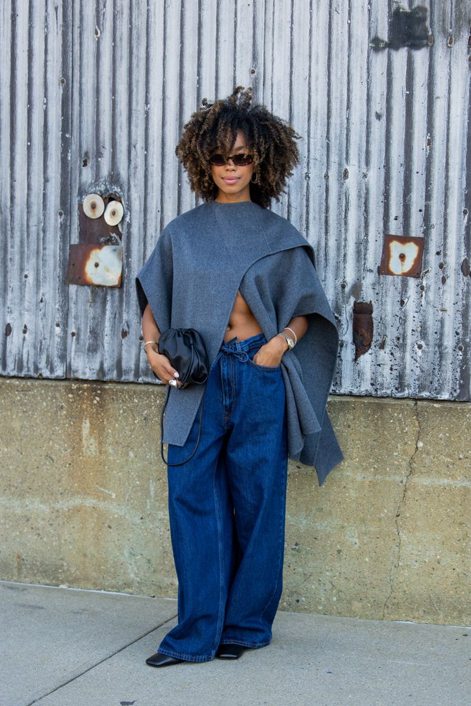 Amaka Hamelijnck wears grey wrapped cape, wide leg denim jeans, black bag outside Cos during New York Fashion Week on September 10, 2024 in New York City