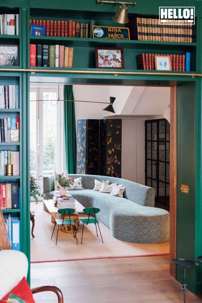 Nina Litchfield's green library at Notting Hill home. A view through a doorway flanked by green bookshelves into a cozy living room with a curved sofa, patterned folding screen, and a small table and chairs.