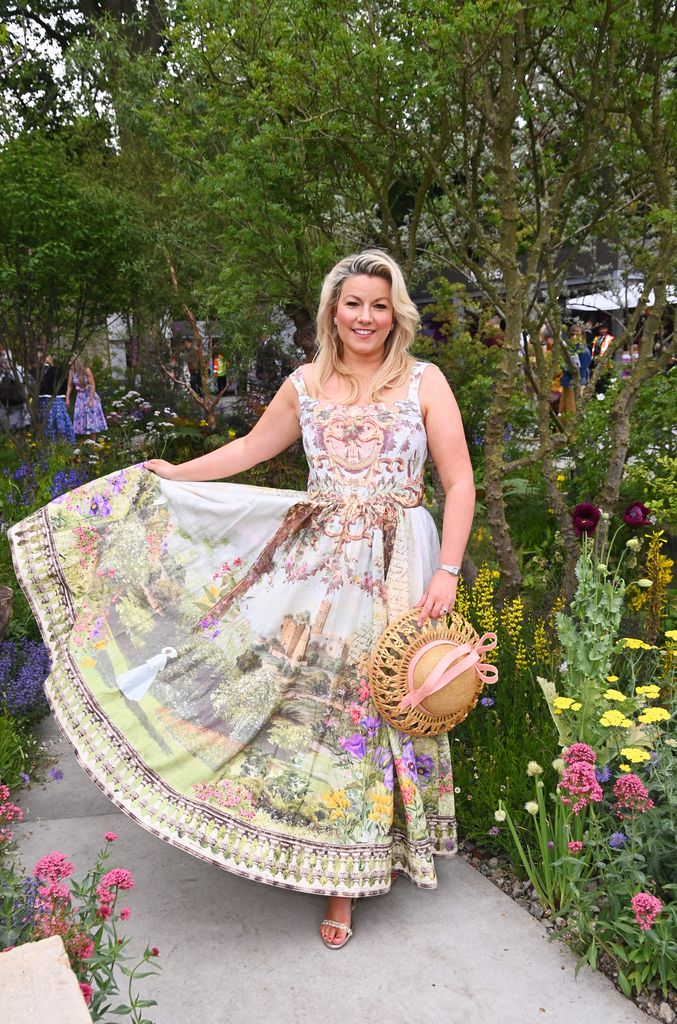 Natalie Rushdie poses in a full-length printed dress with a castle garden motif, holding a straw hat among vibrant flower beds.