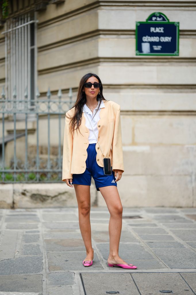 Gabriella Berdugo wears black sunglasses, a white shirt a beige wavy order jacket, gold and silver chain necklaces, a white pearls phone case crossbody bag, blue sport shorts from Adidas, pink shiny leather ballerinas from Roger Vivier,