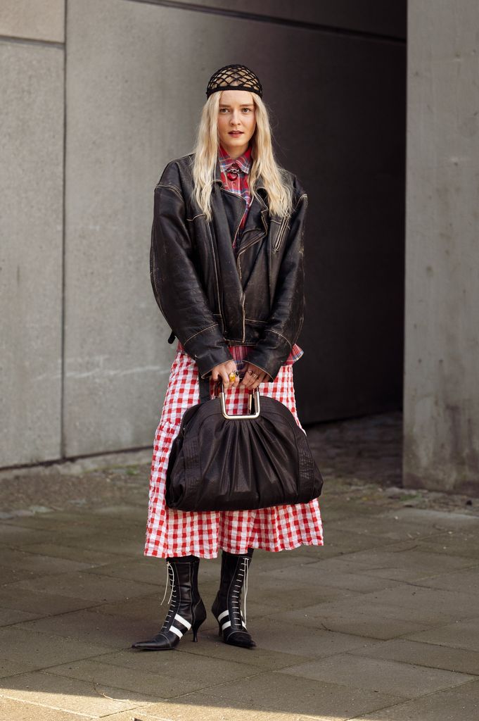 A guest wears red and white checkered dress, black and white pointy shoes, black bag, brown leather jacket, knitted hat outside A. Roege Hove show during the Copenhagen Fashion Week Autumn/Winter 2025 