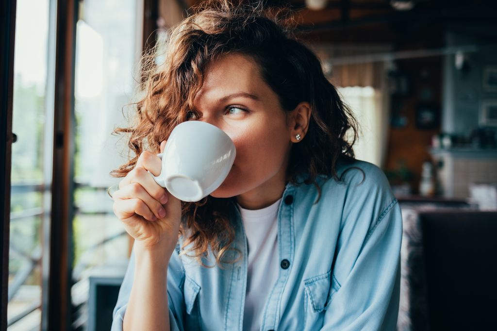 Portrait of happy beautiful young woman wearing blue shirt drinking coffee sitting at table in restaurant looking out the window on summer day.