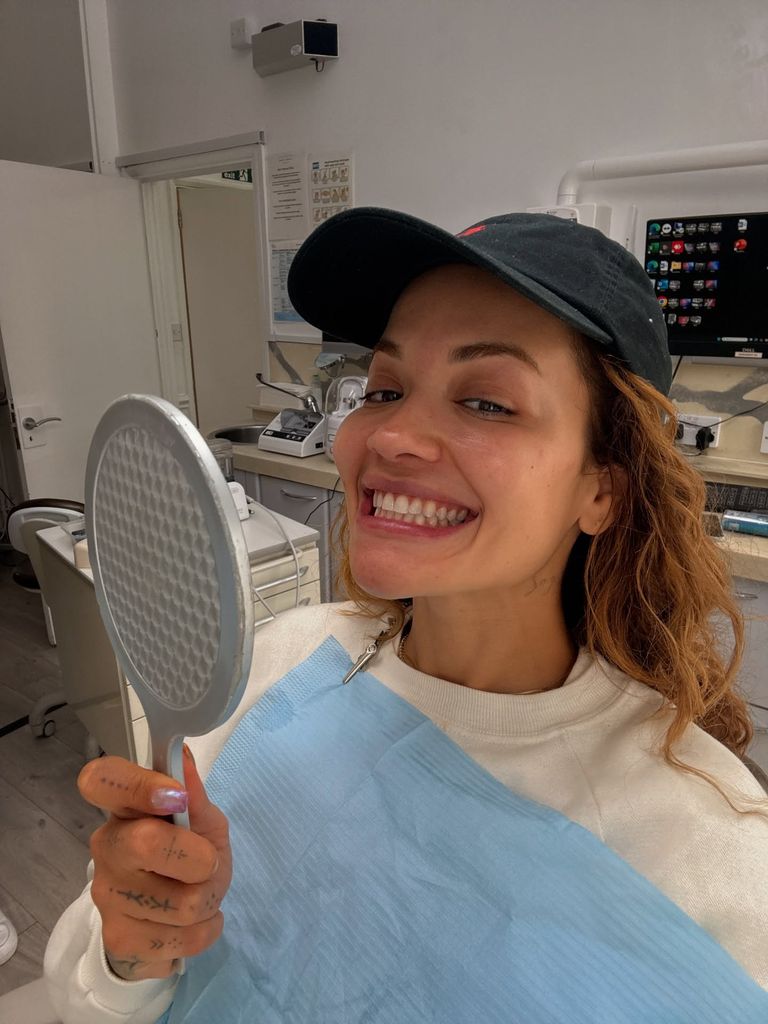 Rita Ora smiles at the camera in a dentist's office, holding a mirror up to her face and wearing a black baseball cap