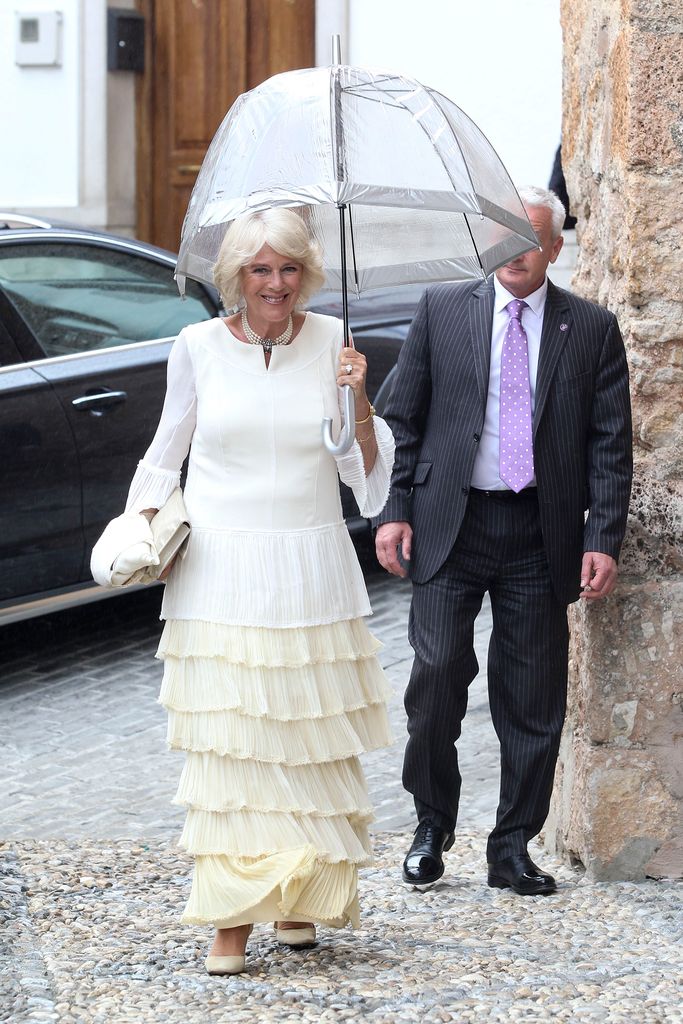 Queen Camilla in a white ruffled dress carrying a clear umbrella