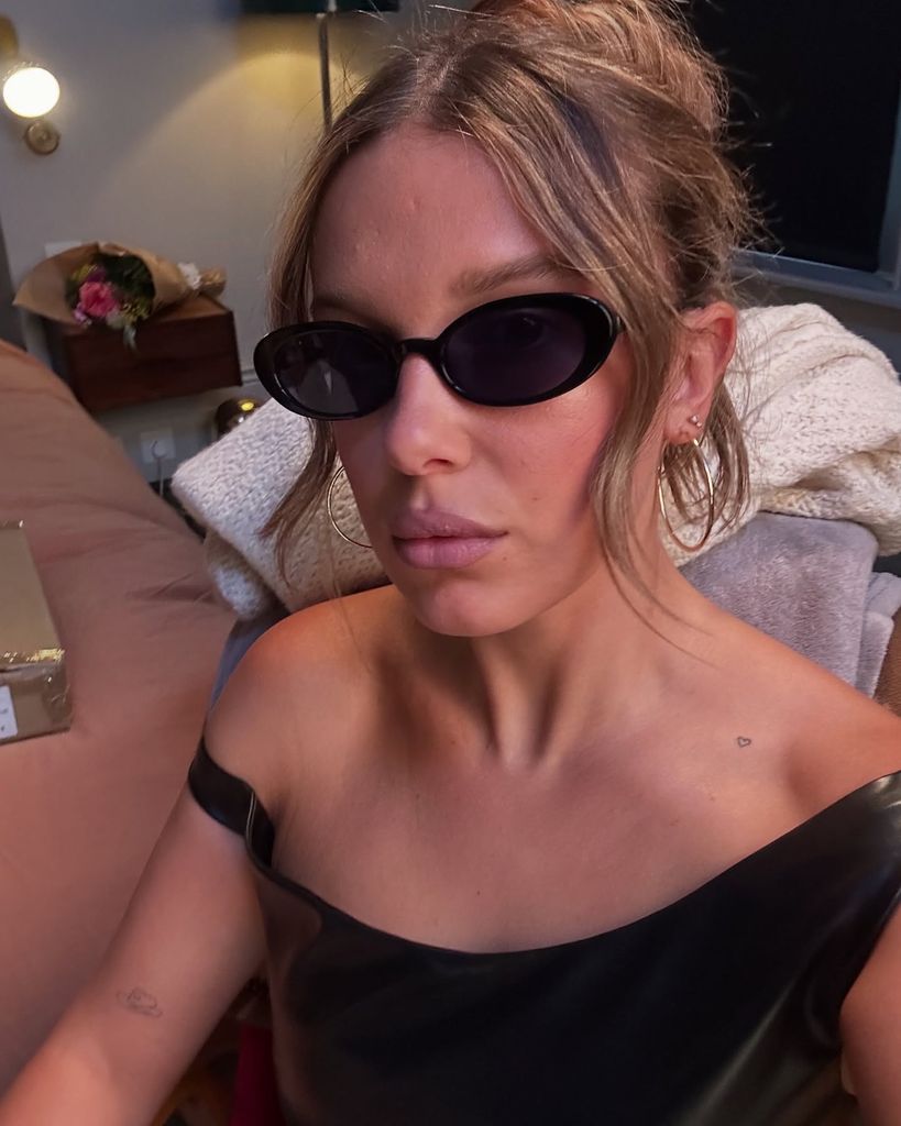 In this close-up selfie, Millie Bobby Brown is seated indoors, facing the camera with a pouty expression. She is wearing sleek, oval-shaped black sunglasses reminiscent of early 2000s fashion and a one-shoulder black leather-look top, which reveals minimalistic tattoos—a small outline of a heart near her collarbone and the Vivienne Westwood orb on her forearm. Her light brown hair is styled up with a few soft tendrils framing her face. Gold hoop earrings complete the Y2K-inspired look. The background includes a pink bedspread, a bouquet of flowers on a nightstand, and a gold-wrapped gift box, suggesting a celebratory or intimate setting.