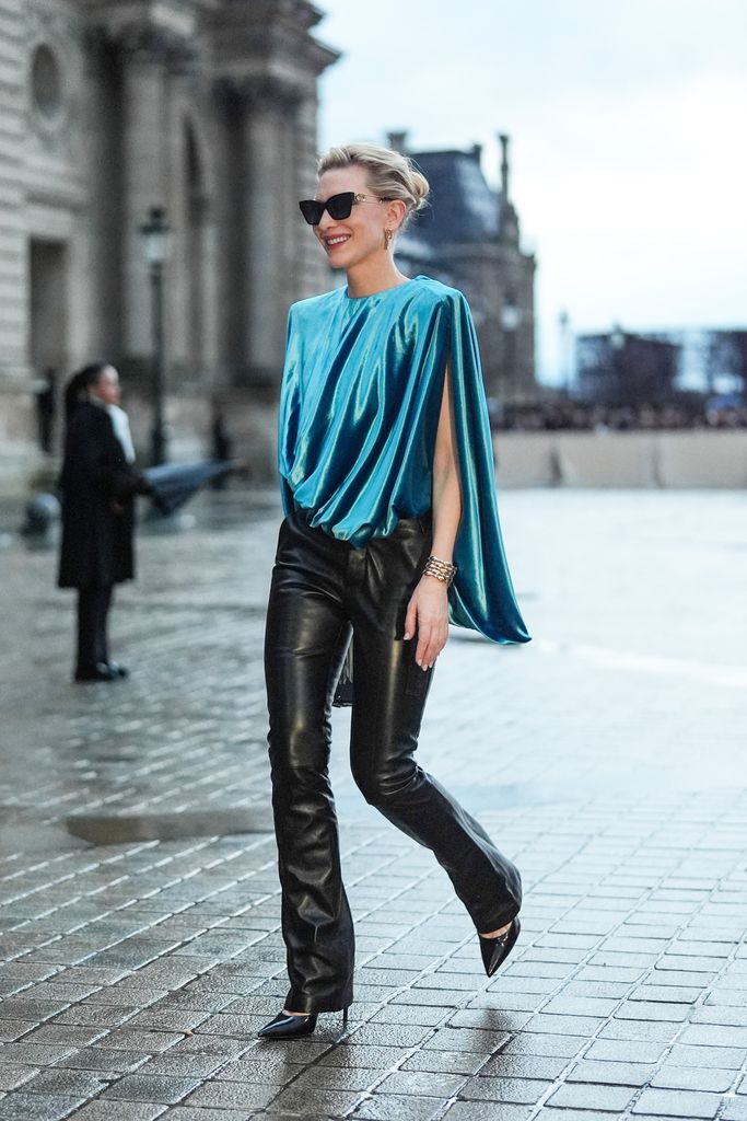 cate walking in a blue turquoise gathered sleeveless lustrous shiny silk top, a bracelet, black leather flared pants
