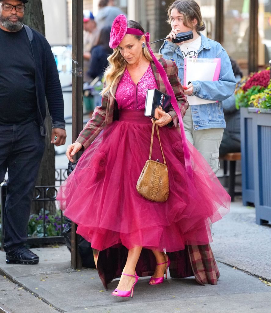 Carrie Bradshaw wears a vibrant pink sequined top with a voluminous magenta tulle skirt. She layers a plaid, oversized coat for contrast. Accessories include a bright pink fascinator, oversized earrings, a tan woven purse, and hot pink strappy heels. She holds a book, exuding confidence and signature bold style.