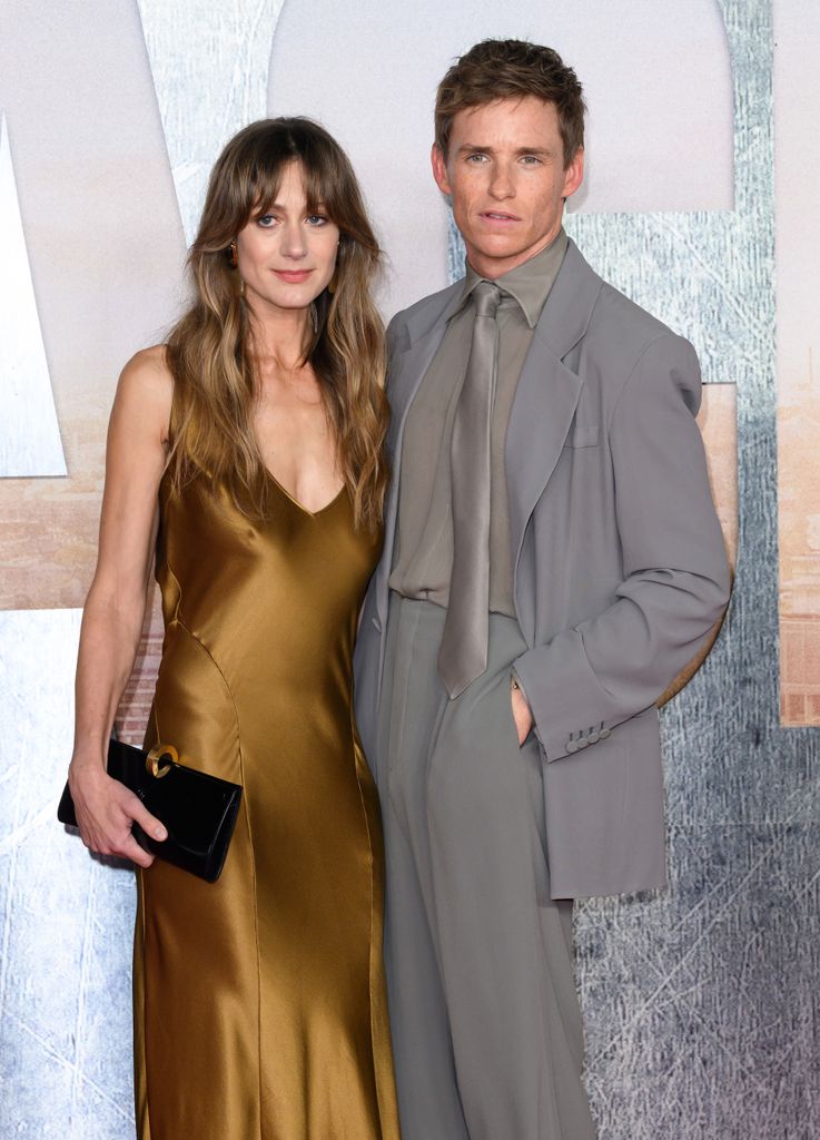 Eddie Redmayne in a grey suit posing next to Hannah Bagshawe in a gold silk dress