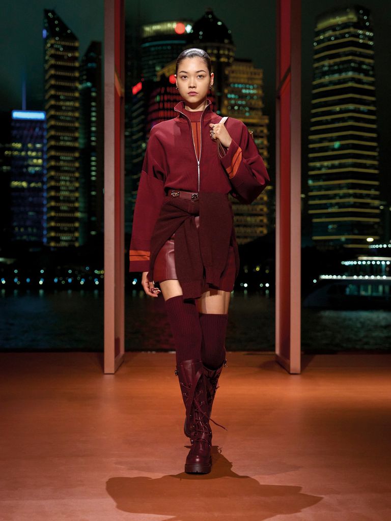 A model walks the runway in a burgundy zip up jacket, mini skirt and knee high boots and socks