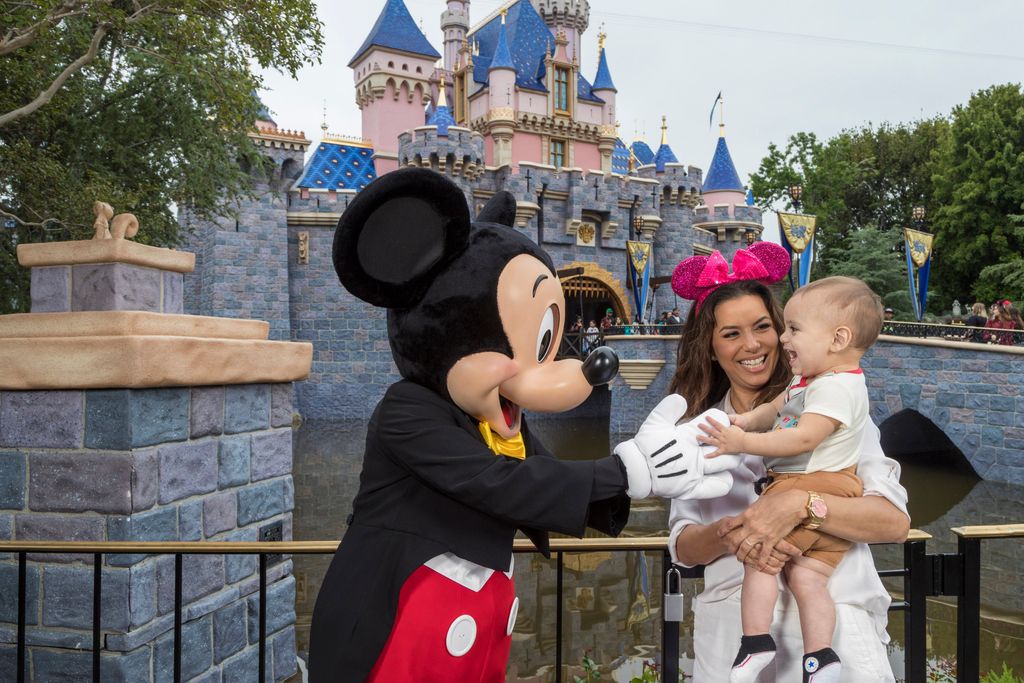 Eva Longoria holding a baby who is being greeted by Mickey Mouse