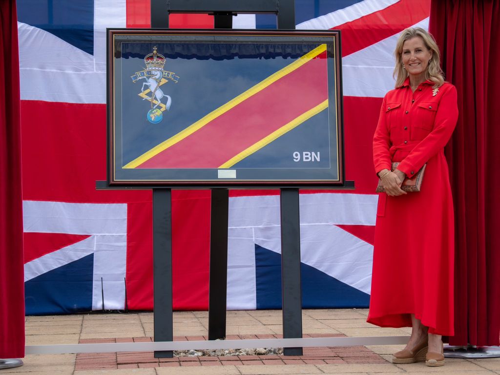 Duchess Sophie wears a red silk dress and stands in front of a large union jack flag