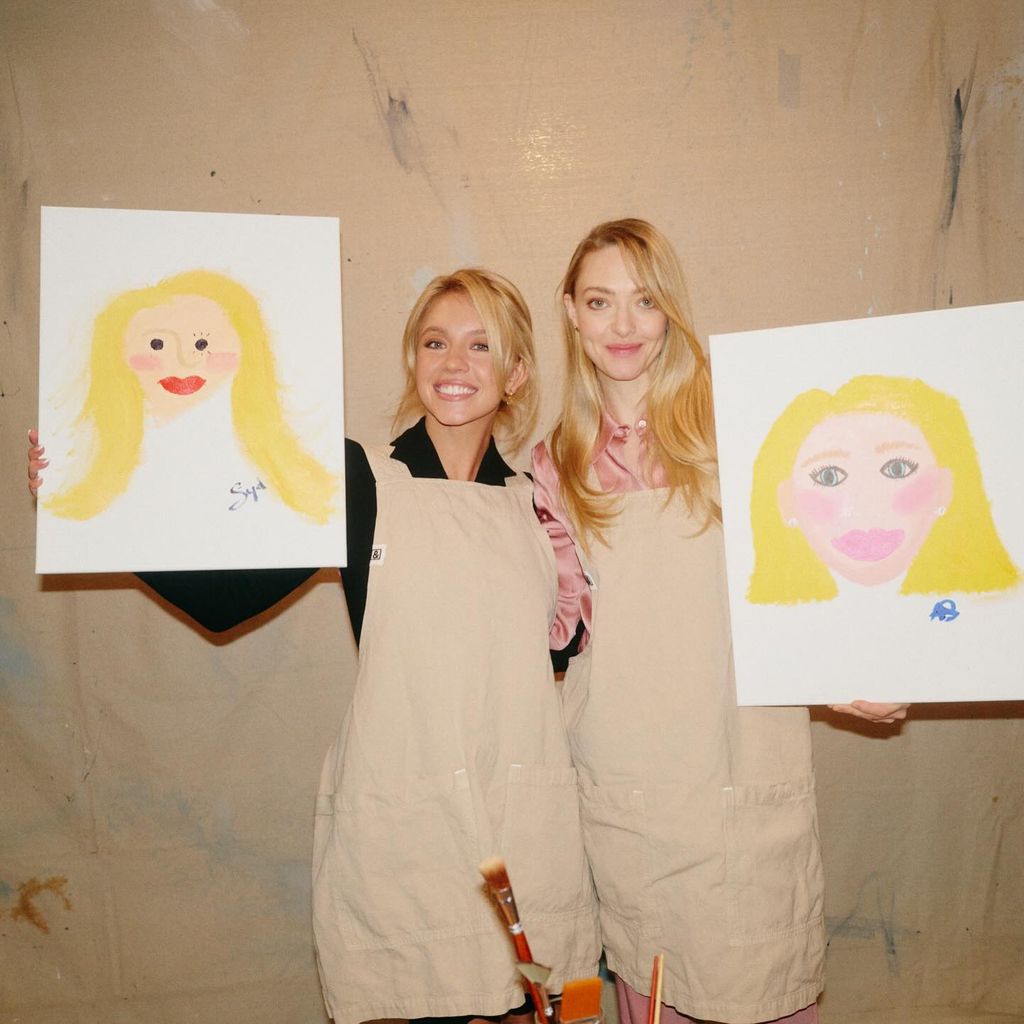 Sydney Sweeney and Amanda Seyfried pose wearing beige aprons, each holding up a picture they have painted of each other. 