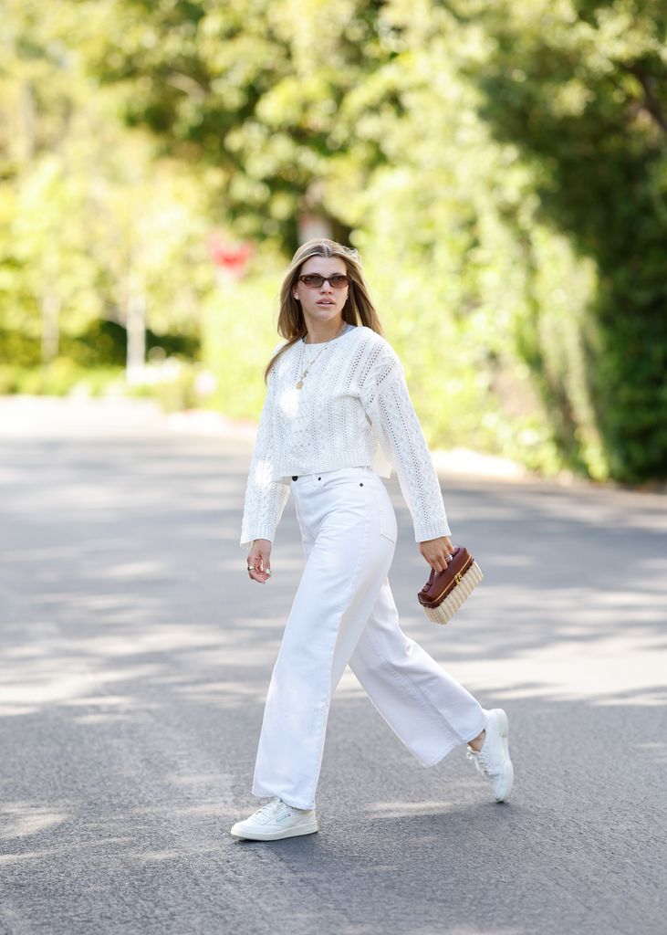 Sofia Richie walks casually under dappled sunlight in a textured ivory sweater, loose white jeans, classic sneakers, gold jewelry, and cat-eye sunglasses, accessorized with a structured mini handbag, embodying relaxed yet polished quiet luxury.